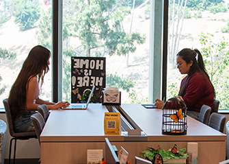 Two students studying together at a table. A Home is Here sign in the background.